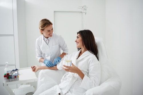 woman sitting in armchair and holding glass of lemon water while doctor in sterile gloves checking IV infusion